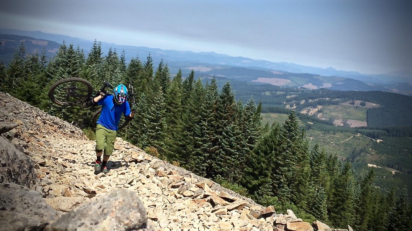 A person wearing a helmet and blue shirt is carrying a mountain bike while climbing a rocky trail surrounded by tall pine trees and scenic mountainous terrain. The sky is partly cloudy, and the landscape features distant hills and valleys. Cold Creek mountain bike trail.