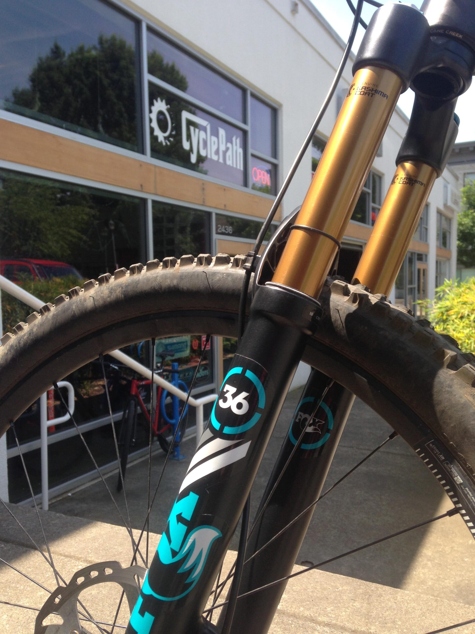 A close-up view of a mountain bike's front suspension forks, showcasing a black and turquoise design with the number "36" prominently displayed. In the background, the exterior of a bike shop called "CyclePath" is visible, featuring large windows and an "OPEN" sign.
