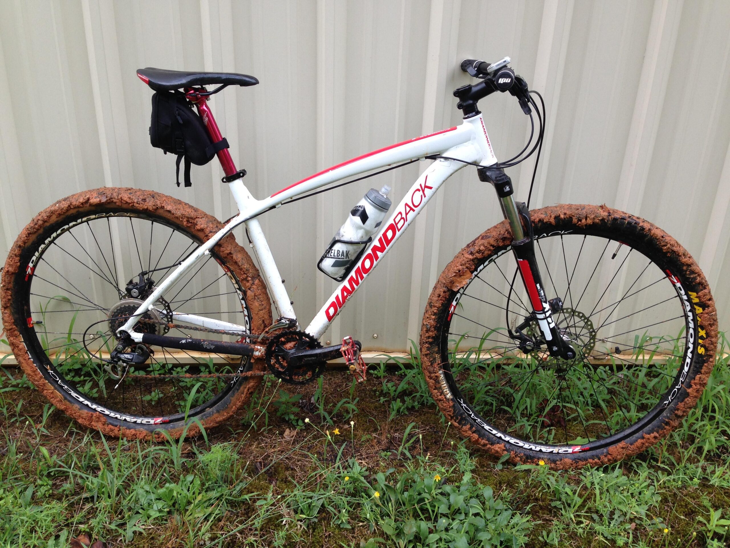 Diamondback Overdrive: A muddy mountain bike resting on grass, featuring a white frame with red accents and brand markings. The tires are caked with mud, indicating recent off-road use. A small black bag is attached to the seat, and there are some green plants and yellow flowers visible in the background next to a light-colored metal wall.