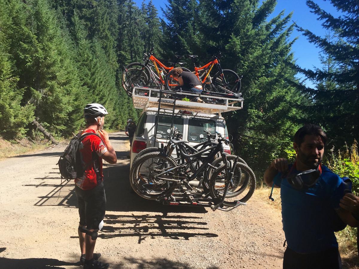 Two mountain bikers preparing for a ride beside a vehicle with a bike rack, loaded with multiple bikes. The scene is set in a forested area with tall green trees and a dirt road. One biker is adjusting his helmet and wearing a red jersey, while the other, dressed in blue, is adjusting his backpack. Alpine Trail mountain bike trail.