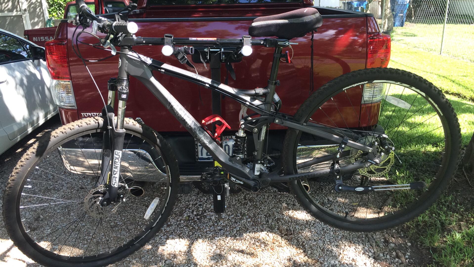 Mongoose MONGOOSE XR-PRO: A black mountain bike mounted on the back of a vehicle, showing its front fork, handlebars, and rear suspension. The background features a red pickup truck and a grassy area with sunlight filtering through trees.