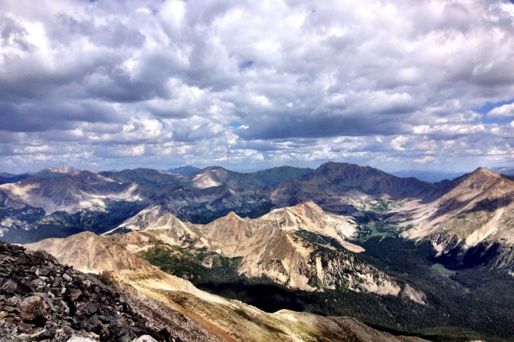 A panoramic view of rugged mountains under a cloudy sky, showcasing a range of peaks and valleys. The landscape features a mix of rocky surfaces, green valleys, and varying shades of brown and gray on the mountainside, creating a dramatic natural scene.