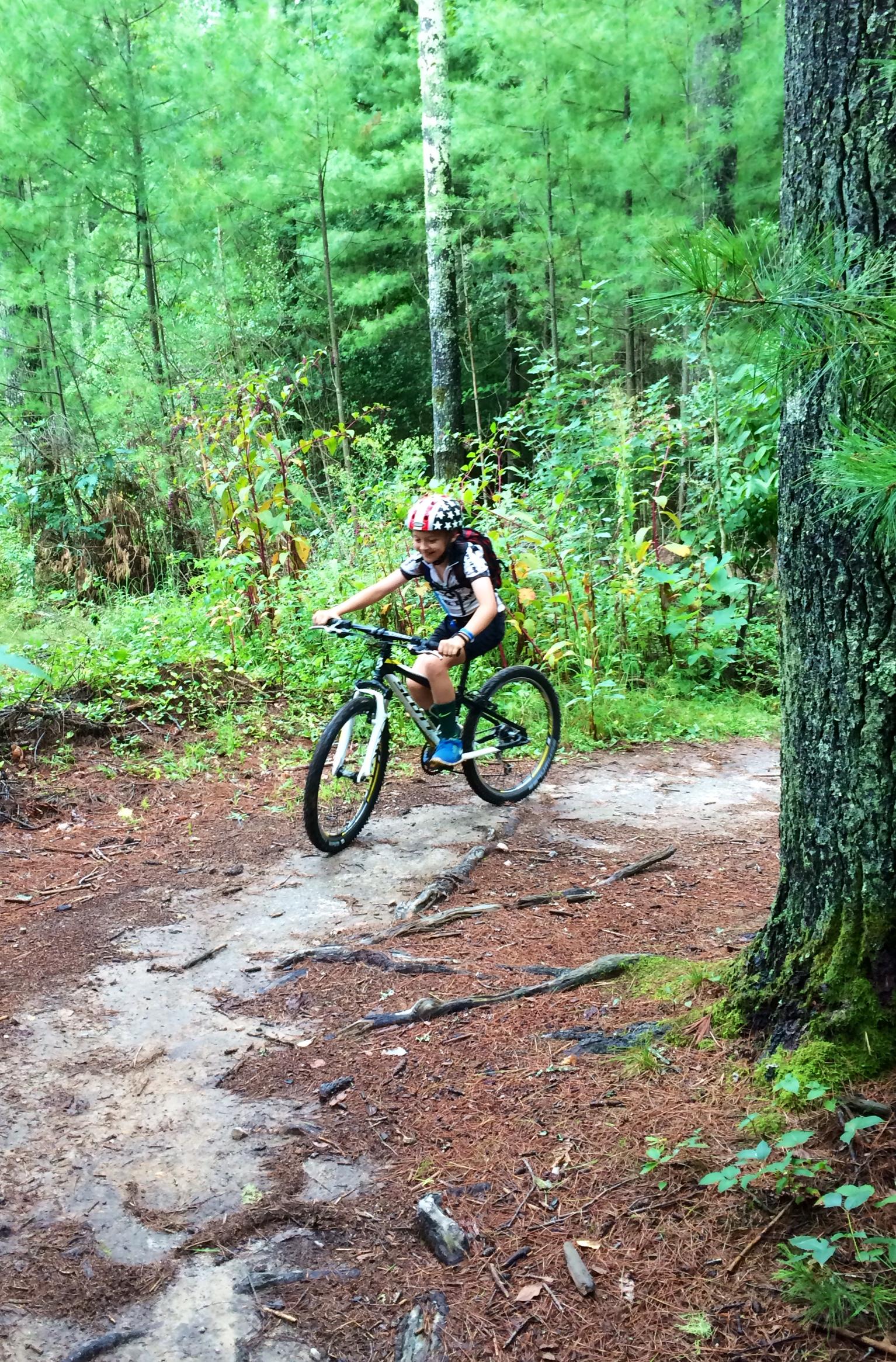 A child riding a mountain bike on a dirt trail through a lush green forest, wearing a helmet and a backpack. The surrounding trees are tall and dense, with patches of colorful foliage. The ground is covered with pine needles and small rocks. DuPont State Forest mountain bike trail.