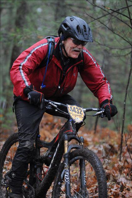 Pivot Mach 429 Carbon: A cyclist in a red jacket and black helmet riding a mountain bike through a wooded area covered with fallen leaves. The cyclist appears focused and determined, with a race number displayed on the bike. The scene is set in a natural, outdoor environment, indicating a challenging terrain.