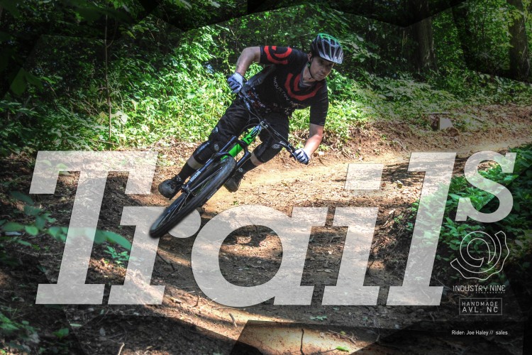 A mountain biker navigating a dirt trail through a lush, green forest, leaning into a turn with determination. The image features overlay text that reads "Trails" and includes branding for Industry Nine, along with the rider's name, Joe Haley.