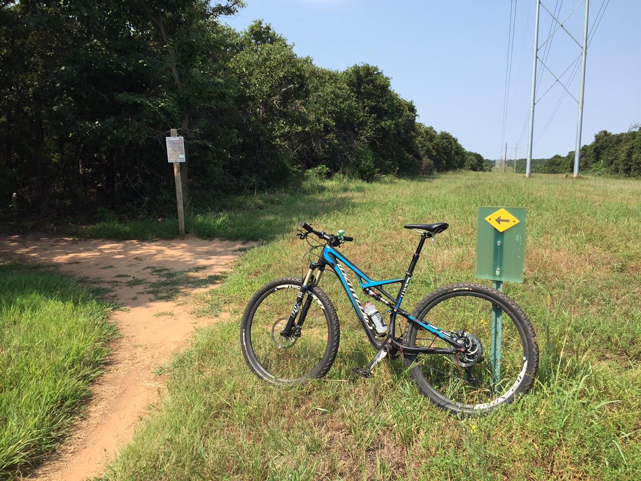 A mountain bike parked on a trail next to a directional sign indicating a left turn. In the background, there are trees and power lines, with a sunny sky overhead. The trail has a sandy path leading into the woods. Lake Stanley Draper mountain bike trail.