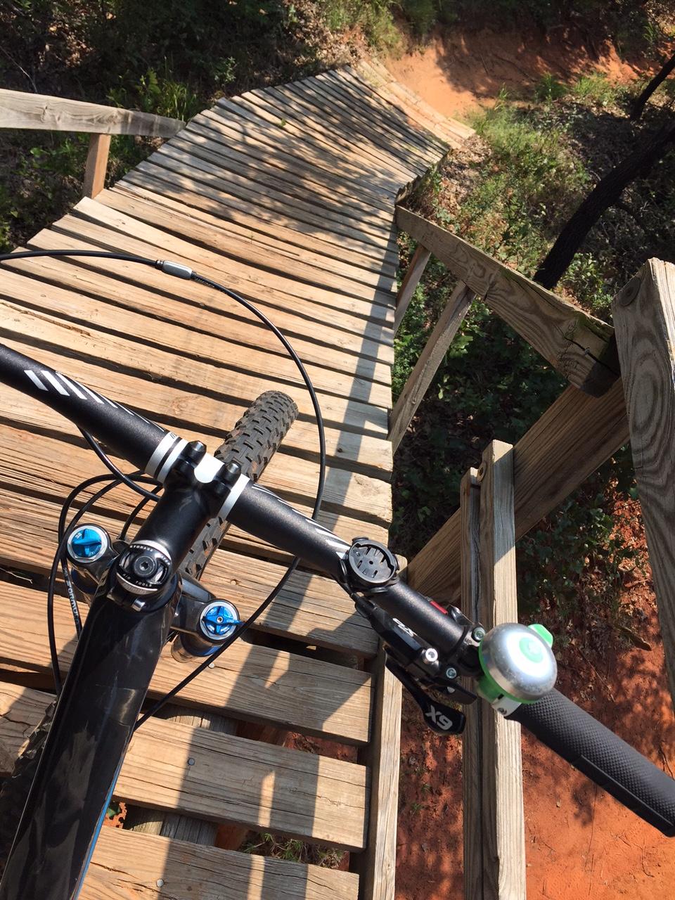 A bird's-eye view of a wooden bike ramp winding downward through a wooded area, with a mountain bike in the foreground. The handlebars, equipped with a bell and gear shifter, are visible, and the ramp descends toward a sandy path surrounded by greenery. Lake Stanley Draper mountain bike trail.