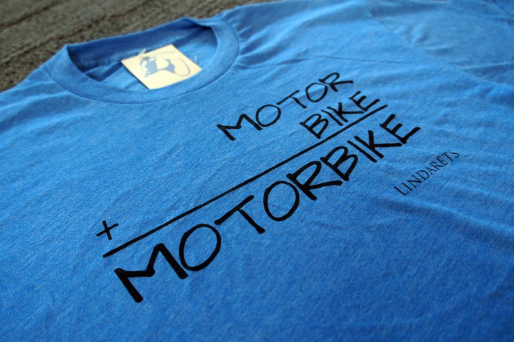 A blue t-shirt laying flat, featuring a graphic design that reads “MOTOR BIKE + MOTORBIKE” in bold black letters, with “LINDARETS” printed in a smaller font beneath the design. The shirt has a classic crew neck style.
