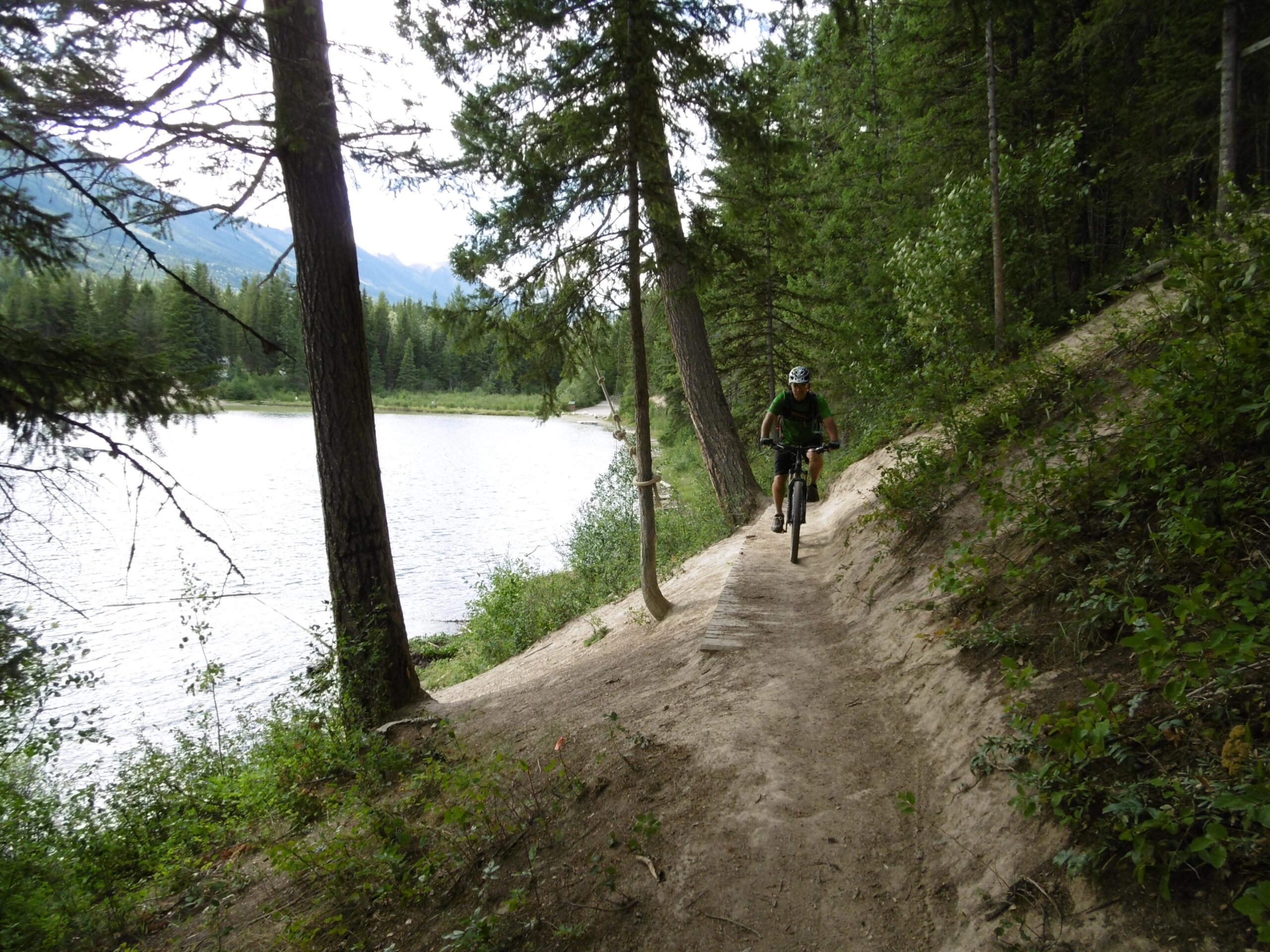A mountain biker rides along a narrow dirt trail beside a serene lake, surrounded by tall trees and lush greenery. The scene captures the beauty of nature, with mountains visible in the background under a cloudy sky. Moonraker Trails mountain bike trail.