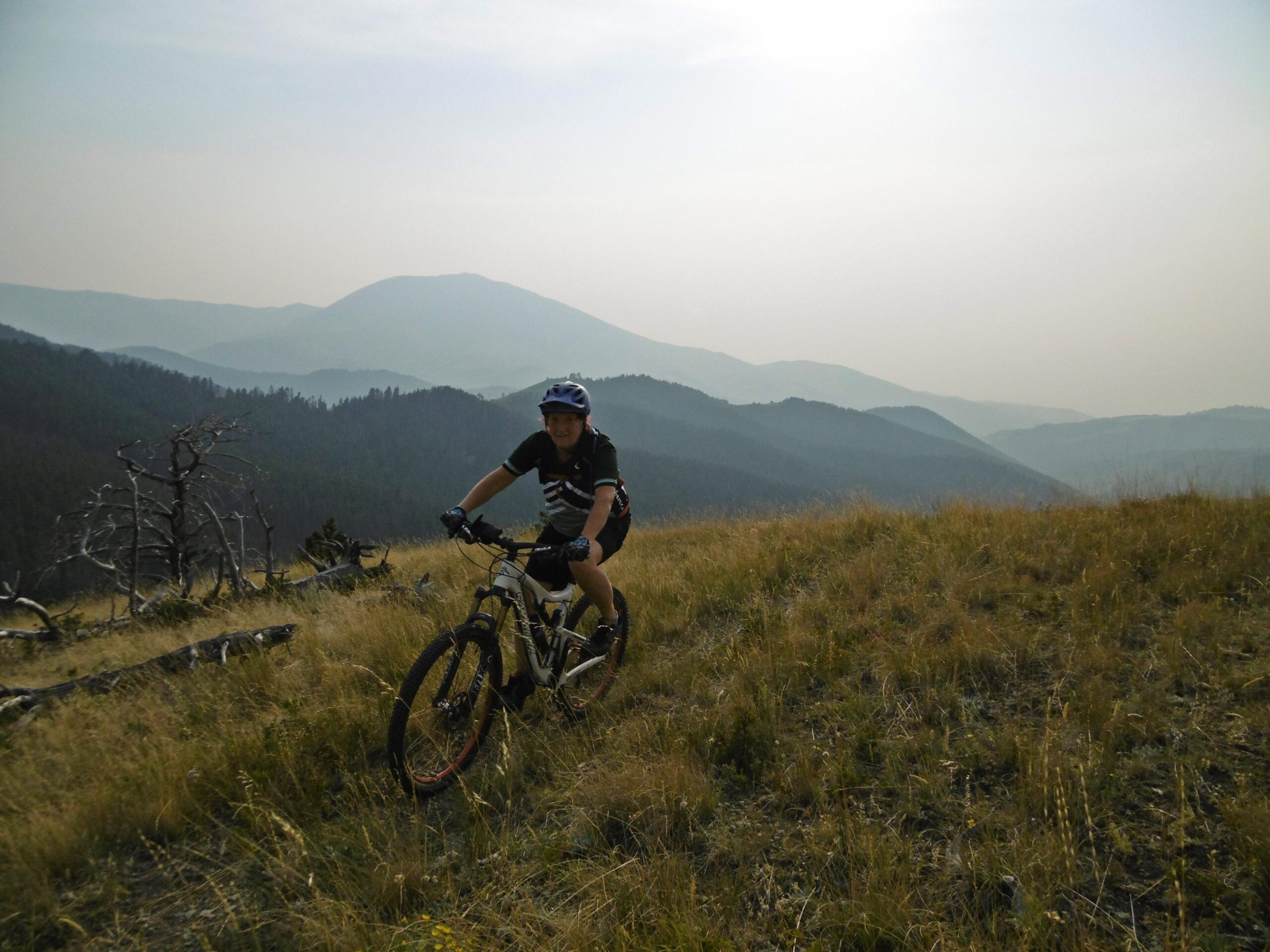 A mountain biker riding through a grassy hillside with distant mountains shrouded in a hazy atmosphere. The biker is wearing a blue helmet and a black shirt with white stripes, enjoying the scenic landscape. Windy Mountain Loop mountain bike trail.