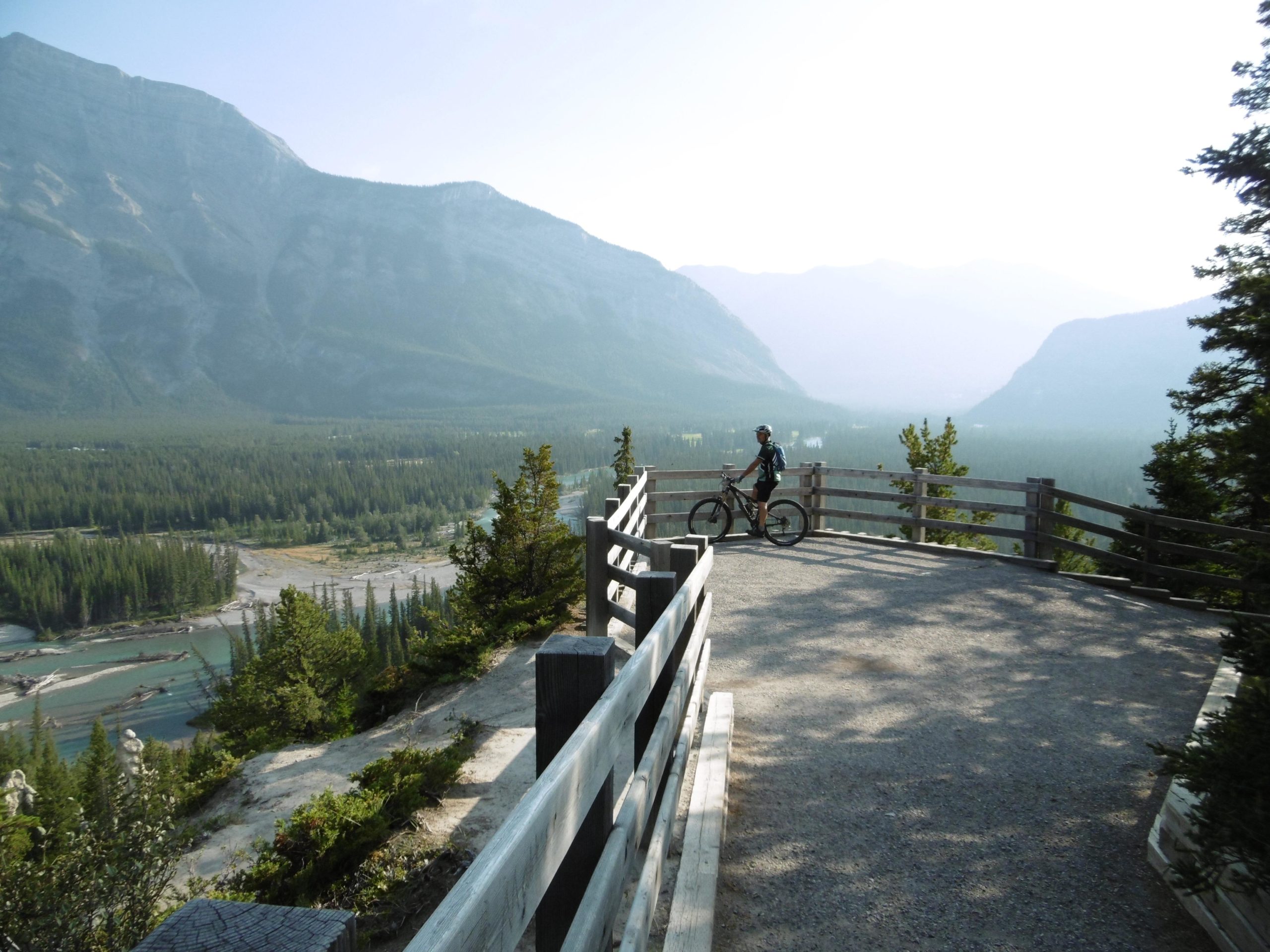 A mountain biker stands on a lookout point, surrounded by towering mountains and lush green forests. The view stretches down to a winding river below, with a hazy atmosphere adding to the picturesque landscape. Wooden guardrails frame the scene, providing safety for those enjoying the stunning scenery. Tunnel Mountain Trail System mountain bike trail.