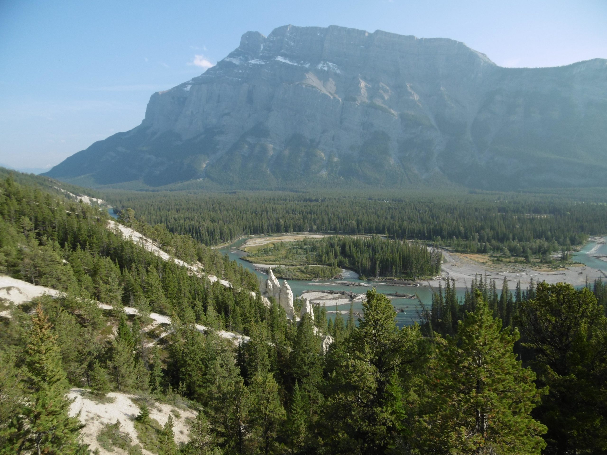 A panoramic view of a mountainous landscape featuring a prominent mountain peak in the background, lush green forests in the foreground, and a winding river flowing through a valley. The sky is clear, with a hint of haze, creating a serene and picturesque natural setting. Tunnel Mountain Trail System mountain bike trail.