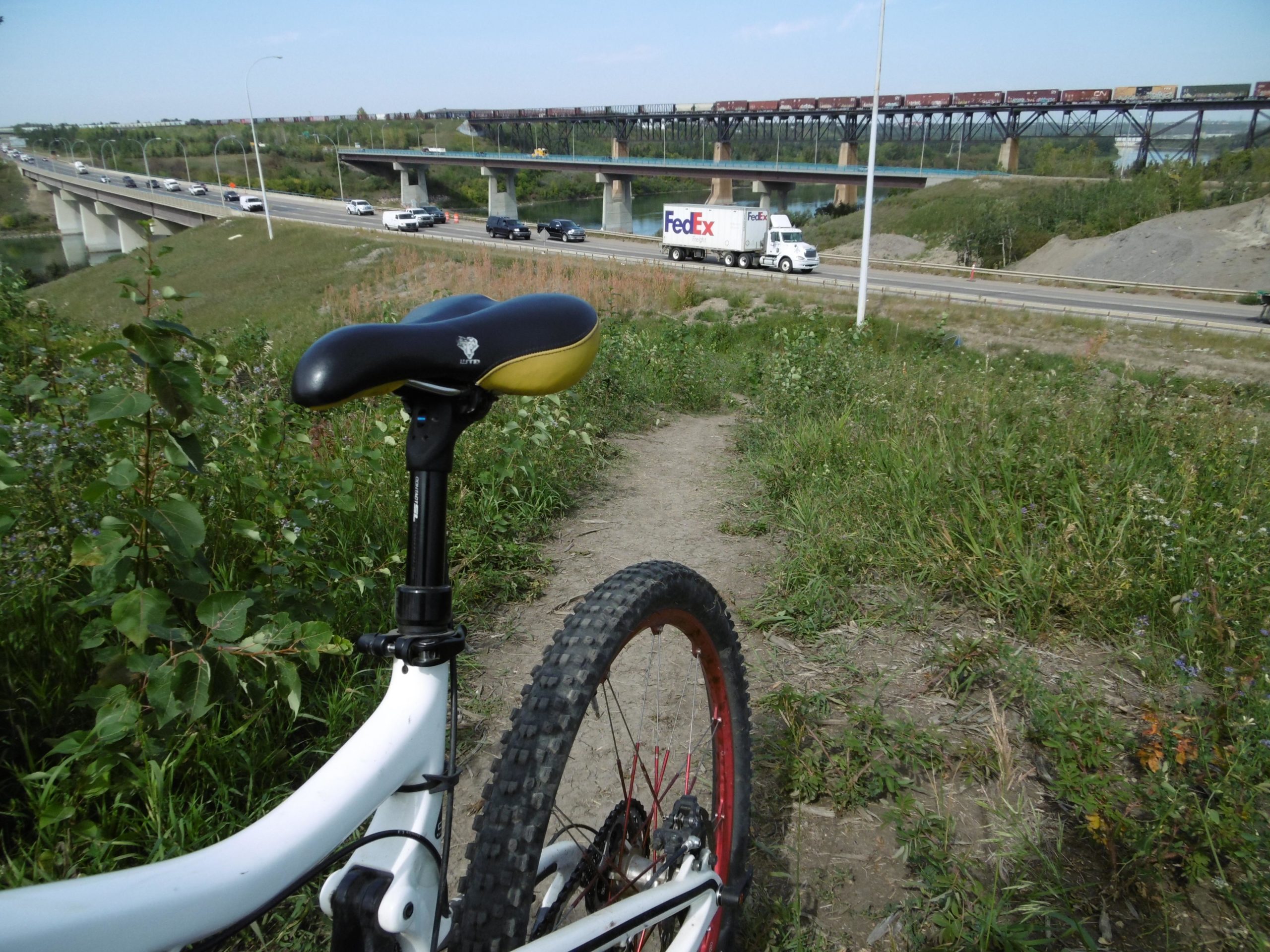 A white mountain bike is positioned on a dirt path, with its saddle and wheel in focus. In the background, a highway is visible with several vehicles, including a truck, while a train travels on a bridge above. Lush greenery surrounds the path, creating a natural environment alongside the urban setting. Strathcona Science Park mountain bike trail.