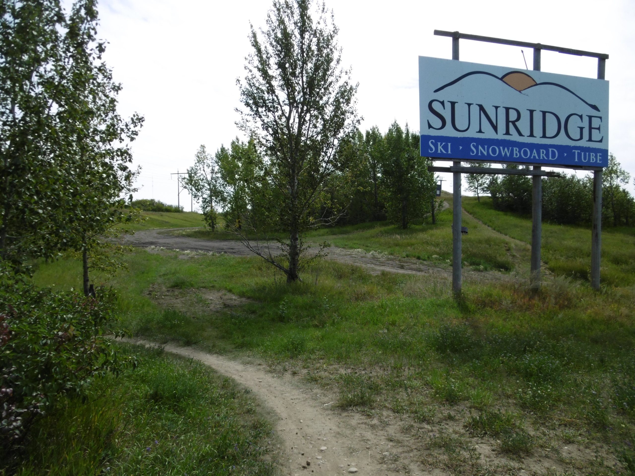 Sign for Sunridge Ski Resort, indicating activities for skiing, snowboarding, and tubing, surrounded by greenery and a pathway leading up a hill. Strathcona Science Park mountain bike trail.