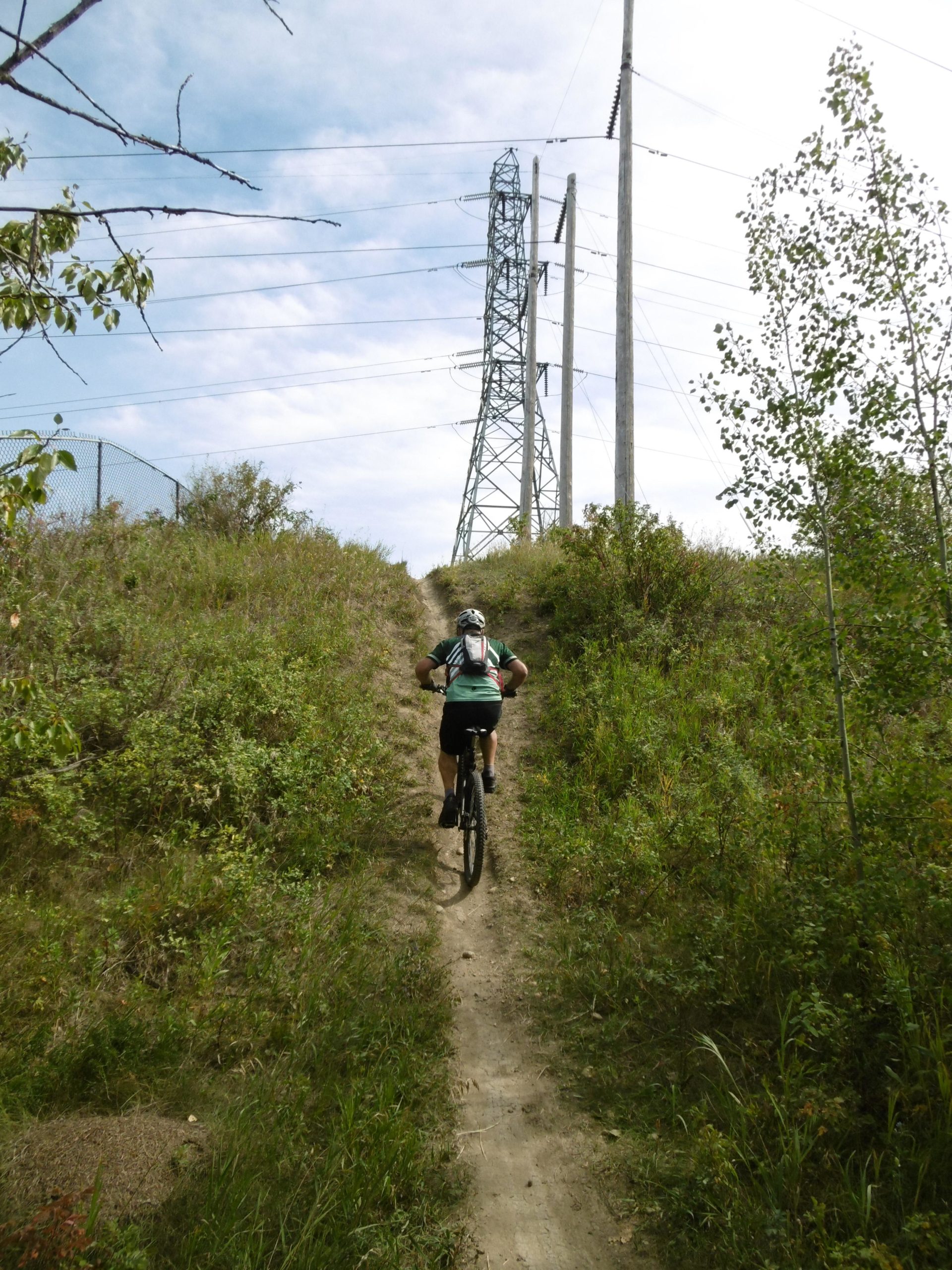 A mountain biker ascends a narrow, grassy trail surrounded by greenery, with a power line and electrical tower visible in the background under a partly cloudy sky. Strathcona Science Park mountain bike trail.