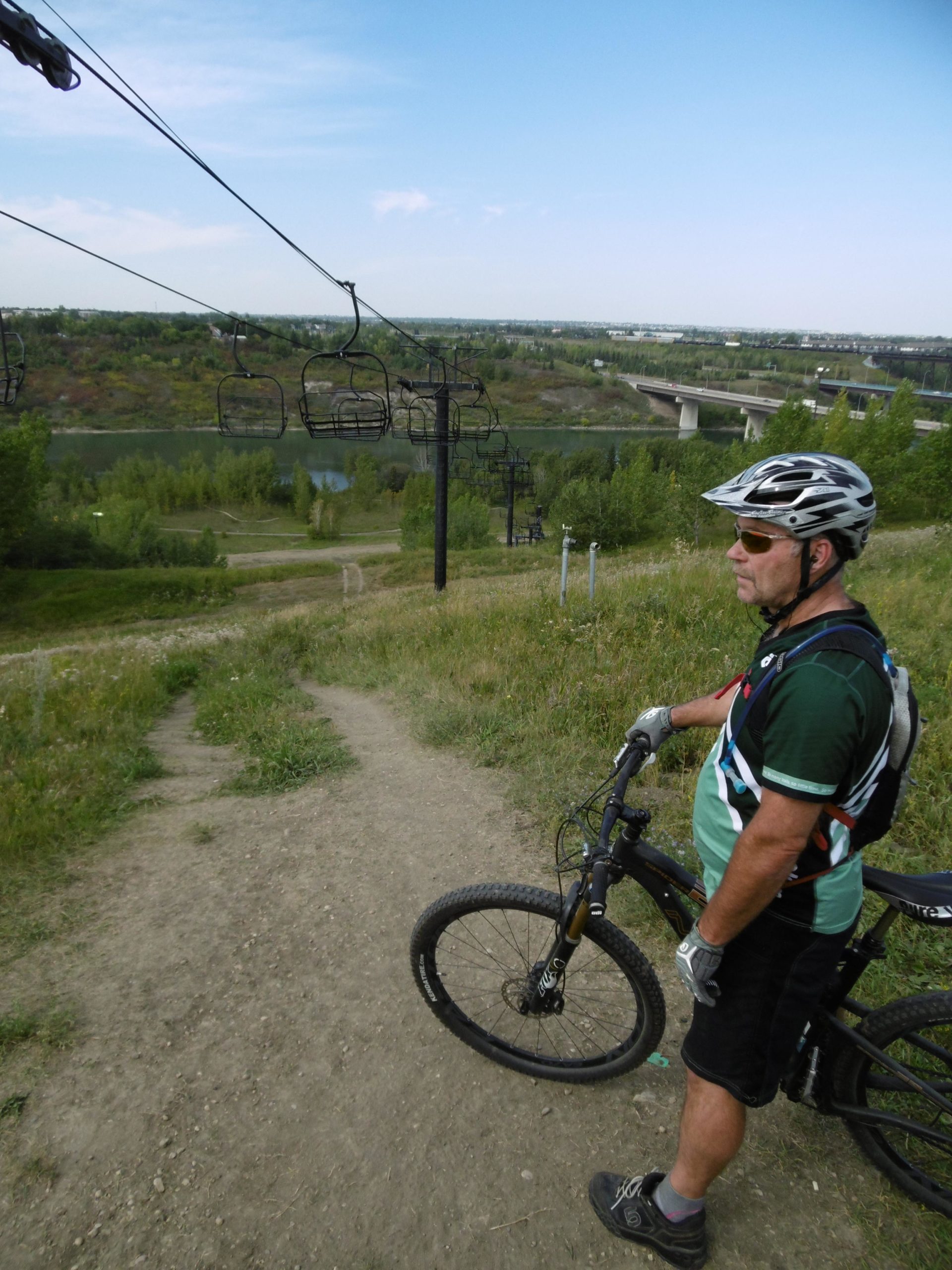 A person wearing a helmet and sunglasses stands next to a mountain bike on a dirt path. In the background, there are ski lift chairs and a scenic view of a landscape with trees and a river. The sky is clear with a few clouds. Strathcona Science Park mountain bike trail.