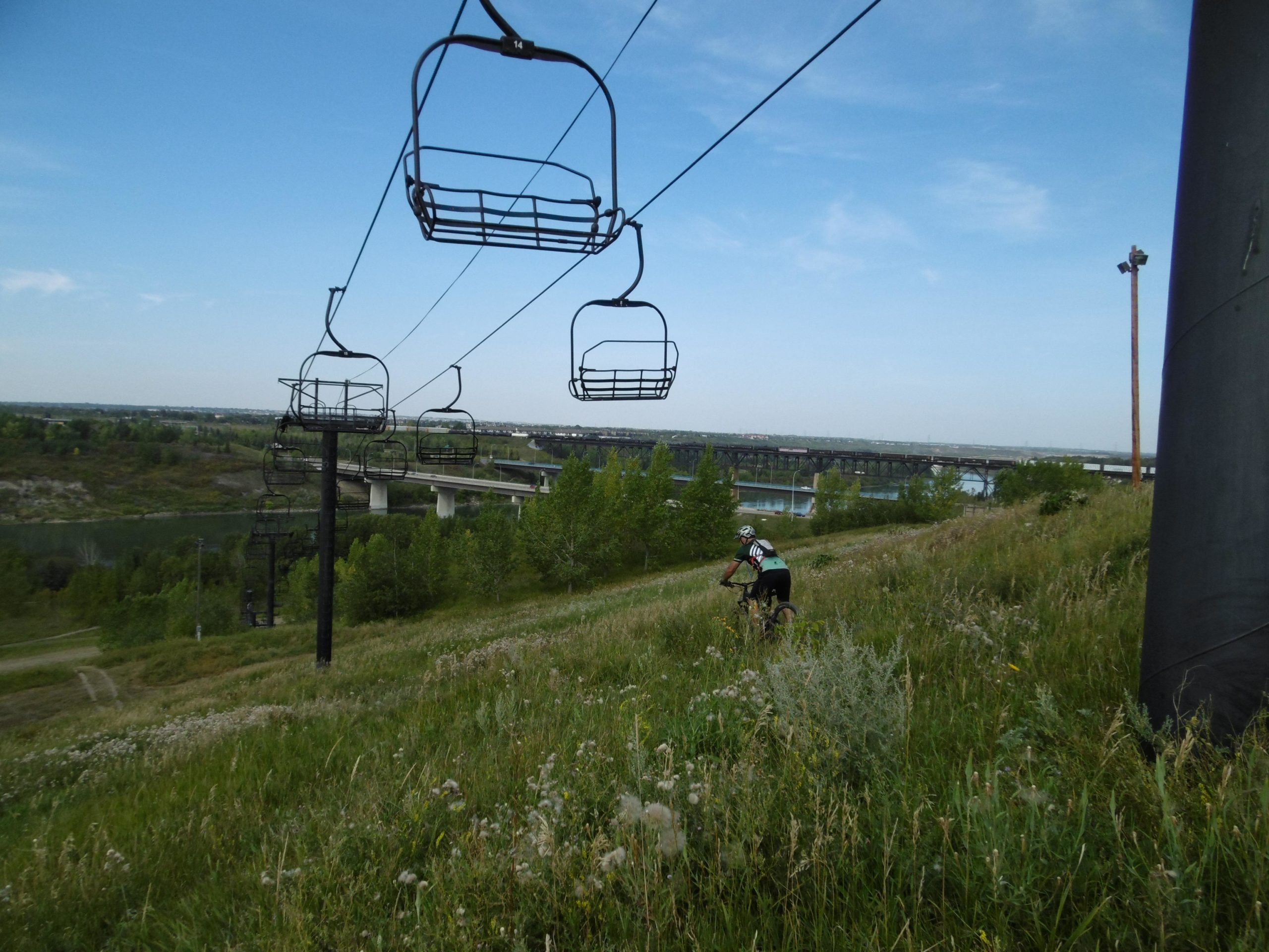 A scenic view of a grassy hillside with a cyclist riding down the slope. In the background, there are chairlifts and a bridge crossing over a river, surrounded by lush greenery under a clear blue sky. Strathcona Science Park mountain bike trail.
