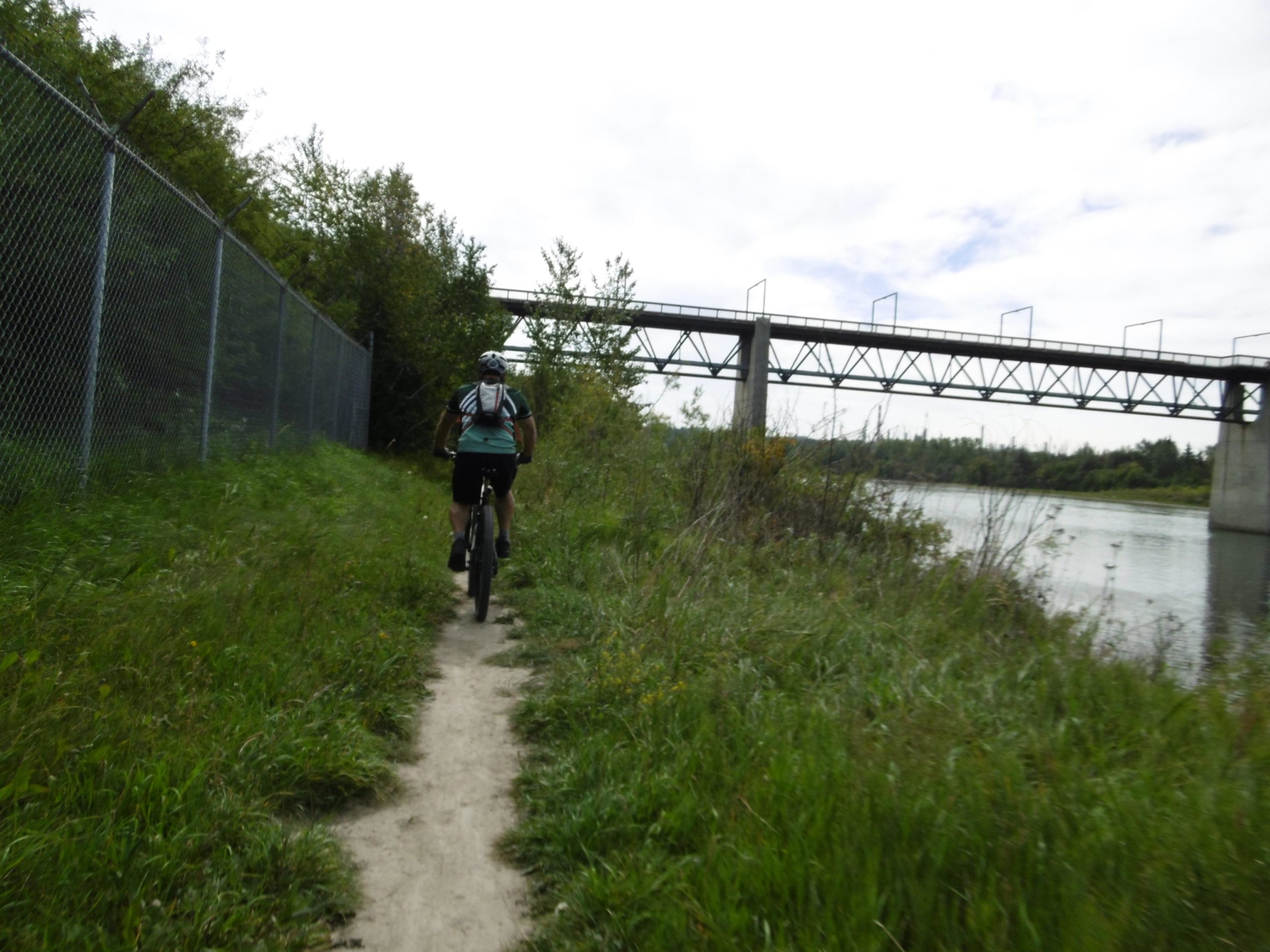 A cyclist riding along a narrow dirt path next to a river, with a chain-link fence on one side and trees in the background. A bridge crosses over the river in the distance under a slightly cloudy sky. Strathcona Science Park mountain bike trail.