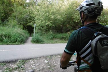 A person wearing a helmet and a biking jersey is standing beside a dirt path that leads into a green, wooded area. The individual is facing the path while holding a bicycle, with a paved road visible in front of them and a grassy landscape surrounding the scene. Mill Creek Ravine (north) mountain bike trail.