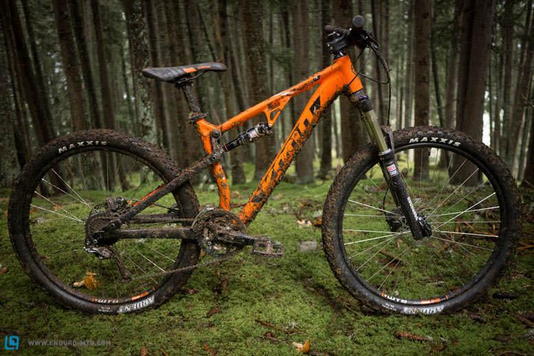 Kona Precept: A muddy orange mountain bike rests on green moss in a dense forest, surrounded by tall trees. The bike shows signs of recent use, with dirt covering the frame and tires, highlighting its adventure-ready design.