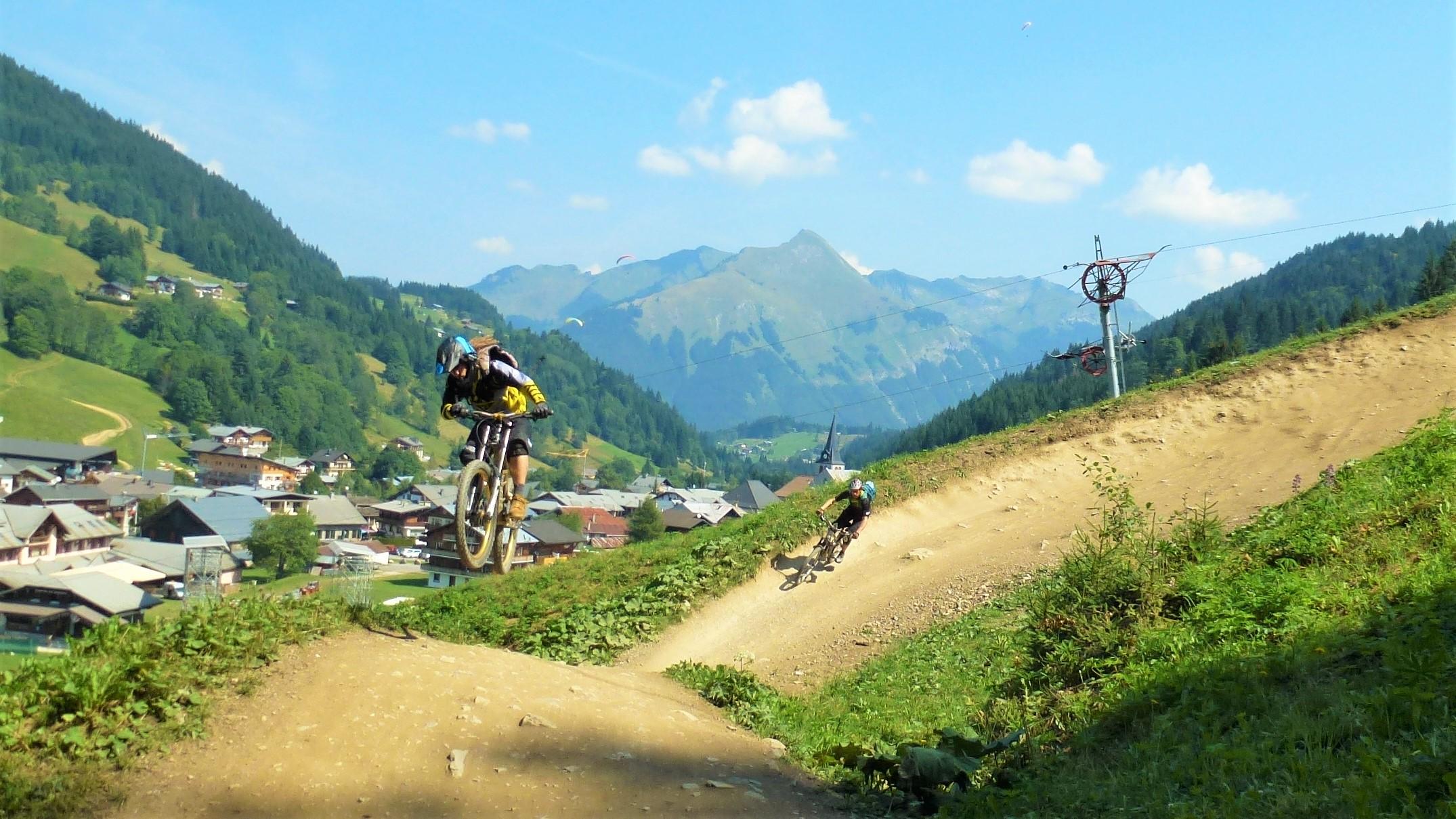 Two mountain bikers riding on a dirt track in a mountainous landscape. One rider is airborne while jumping, and the other is navigating a turn. The background features lush green hills and a small village, with scenic mountains and a clear blue sky above. Chavannes mountain bike trail.