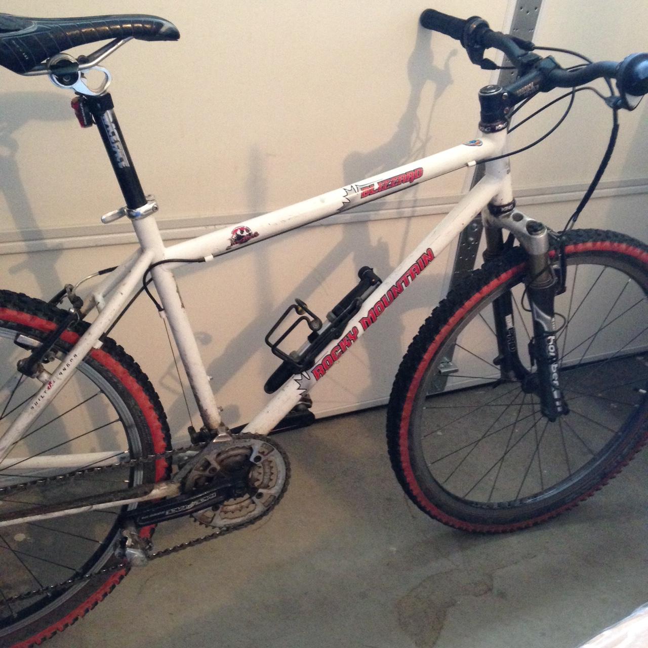 Rocky Mountain Blizzard: Alt text: A white Rocky Mountain mountain bike with red accents is parked on a concrete floor. The bike features a metal frame, thick tires with red rubber, and a black seat. A water bottle holder is attached to the frame. The background shows a garage setting.