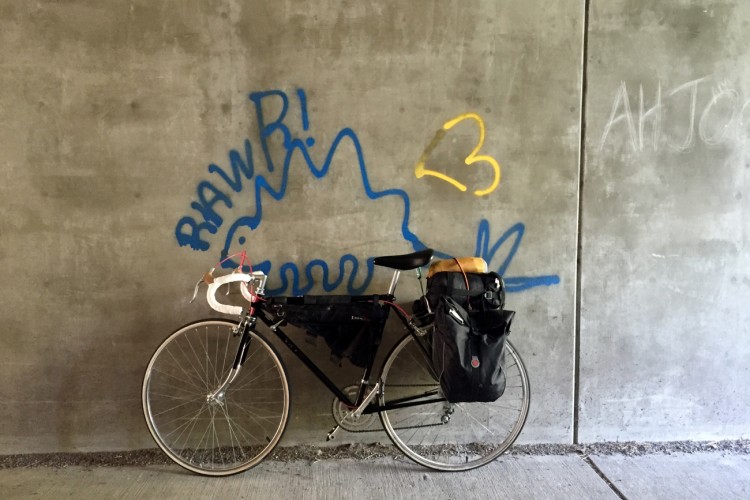 A black bicycle with a decorative handlebar and two side bags leans against a concrete wall. The wall features colorful graffiti, including blue and yellow text and shapes. The setting appears to be an urban environment, possibly a bike path or alleyway.