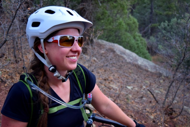 A smiling woman wearing a white helmet and sunglasses, sitting on a mountain bike in a forested area. She has a braided hairstyle and is equipped with a hydration pack. The background features greenery and rocky terrain, suggesting an outdoor biking adventure.
