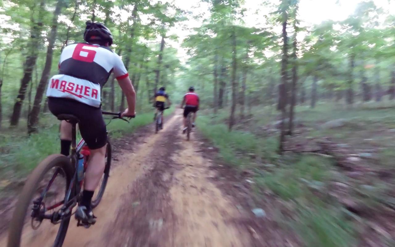Three mountain bikers ride along a dirt trail in a wooded area, surrounded by green trees and foliage. The lead cyclist wears a jersey with the number 6 and the word "MASHER" on it, while the others are in colorful jerseys, reflecting the camaraderie of the group as they navigate the terrain. Oak Mountain State Park mountain bike trail.