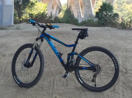 Giant Stance 27.5 2: A blue mountain bike is positioned on a dirt surface, with a backdrop of green foliage and a concrete structure partially visible above. The bike features thick tires and front suspension, indicating it is designed for off-road use.