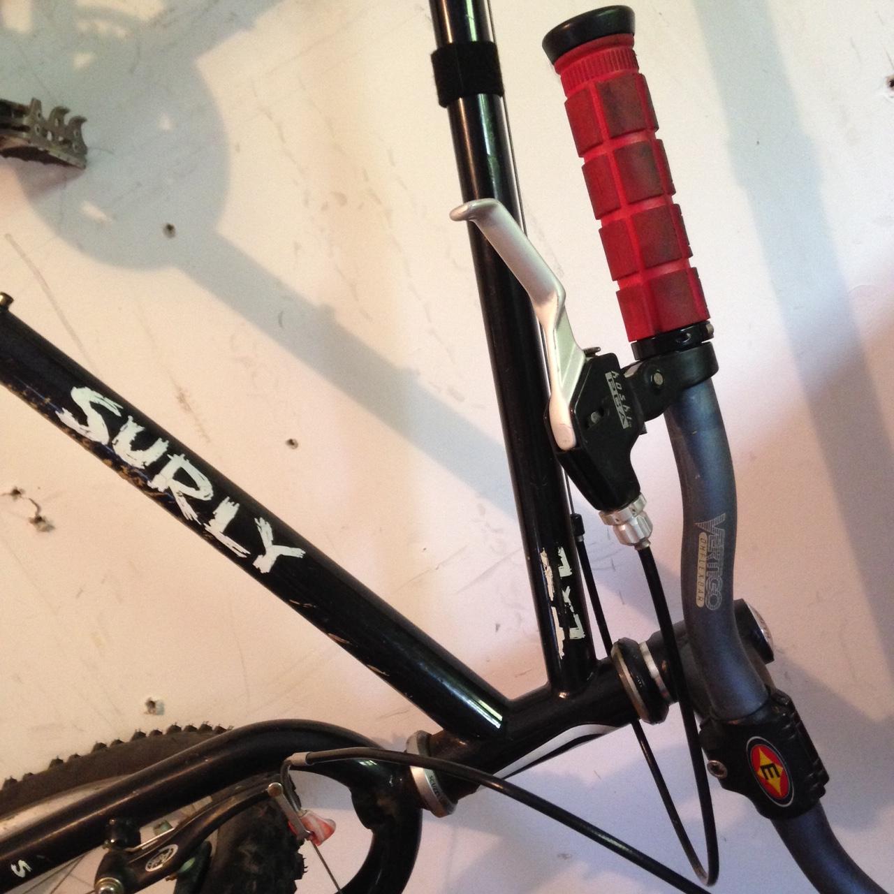 Surly 1x1: Close-up image of a mountain bike frame featuring the brand name "Surly" prominently displayed. The image shows the bike's handlebar with a red rubber grip and a silver brake lever. Various components of the bike are visible, including part of the frame and brake cable. The background is a plain wall with some visible markings.