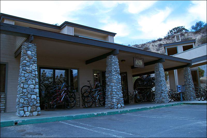 Bicycle shop exterior featuring a covered entryway supported by stone pillars, with several bicycles parked outside and a clear blue sky in the background.
