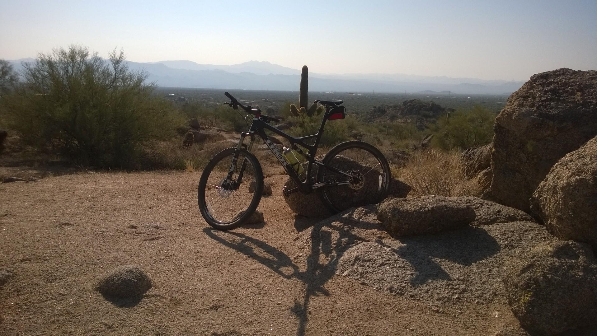 An off-road bicycle leaning against a rock in a desert landscape with cacti, shrubs, and distant mountains under a clear sky. The ground is sandy with a few scattered rocks. Brown's Ranch to Granite Mountain mountain bike trail.