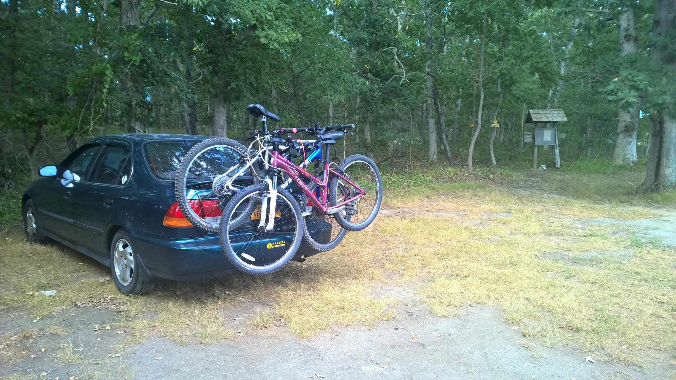 A blue car parked on a gravel area, with two bicycles secured on a bike rack attached to the back. The surrounding area features grass and trees, suggesting a natural outdoor setting. In the background, there is a wooden information kiosk partially visible. Calverton Trail mountain bike trail.