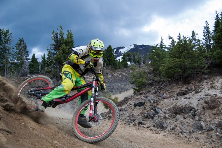 A mountain biker in bright yellow and black gear leans into a turn on a dirt trail surrounded by trees and rocky terrain, with snow-capped mountains visible in the background under a cloudy sky. Dust is kicked up from the rear tire as the rider navigates the trail.