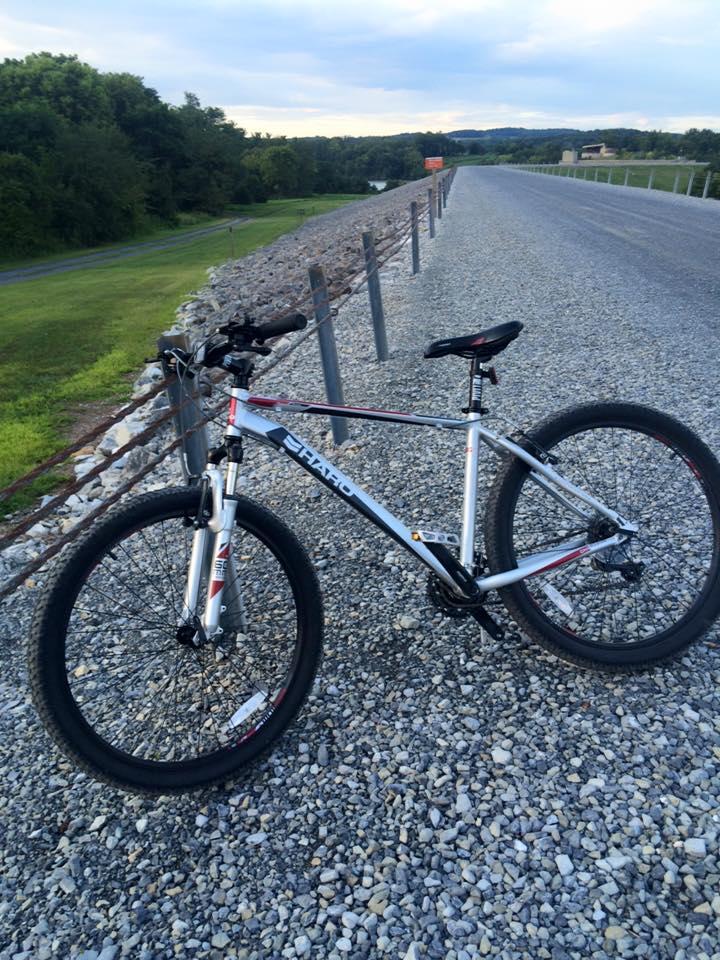 A silver mountain bike parked on a gravel path, with a green landscape and trees in the background. The sky is partly cloudy, indicating a pleasant day for outdoor activities. Blue Marsh mountain bike trail.