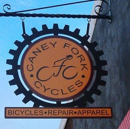Sign for Caney Fork Cycles featuring a bicycle graphic and the text "Bicycles • Repair • Apparel," set against a gear-shaped background.