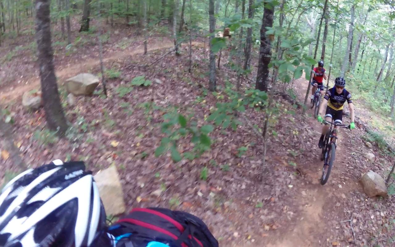 A group of mountain bikers riding on a dirt trail through a wooded area, surrounded by trees and foliage. The path is winding, and there are rocks and leaves scattered on the ground. One biker in the foreground is wearing a helmet and backpack, while two others follow behind him. Coldwater Mountain mountain bike trail.