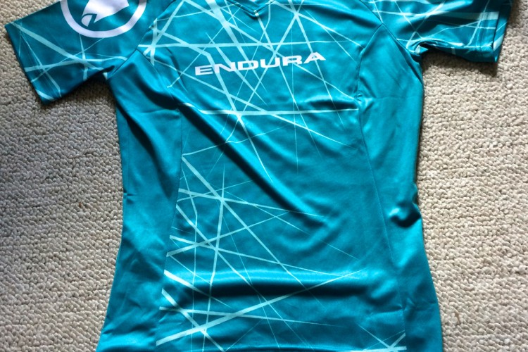 A teal cycling jersey with a geometric pattern and the brand name "ENDURA" displayed prominently on the front. The shirt features short sleeves and a v-neck design. It is laid flat on a textured, light-colored carpet.
