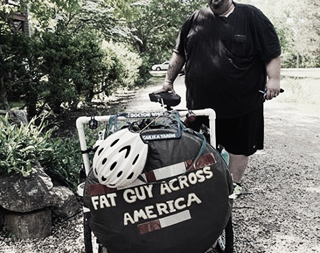 A man stands next to a bicycle with a cart attached, which has a sign that reads "FAT GUY ACROSS AMERICA." He is wearing a black shirt and shorts, and a white bicycle helmet is resting on the cart. The background features greenery and a gravel path.