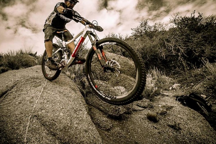 A mountain biker navigating over rocky terrain, showcasing dynamic movement and control. The rider is wearing a helmet and cycling gear, with a bicycle in mid-air as they tackle a challenging obstacle. The background features a rugged landscape with sparse vegetation and a cloudy sky.