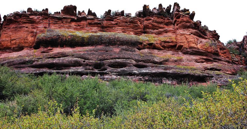 A large, textured rock face with vibrant red and orange hues, featuring layered formations and patches of green moss. The base is surrounded by lush greenery, indicating a rich ecosystem, while the top is rugged with jagged rock formations against a bright sky. Blue Sky mountain bike trail.