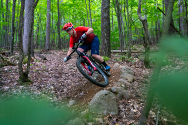 A mountain biker in a red shirt and helmet navigates a dirt path surrounded by green trees, leaning into a turn while riding over rocks and leaves. Hardy Road mountain bike trail.