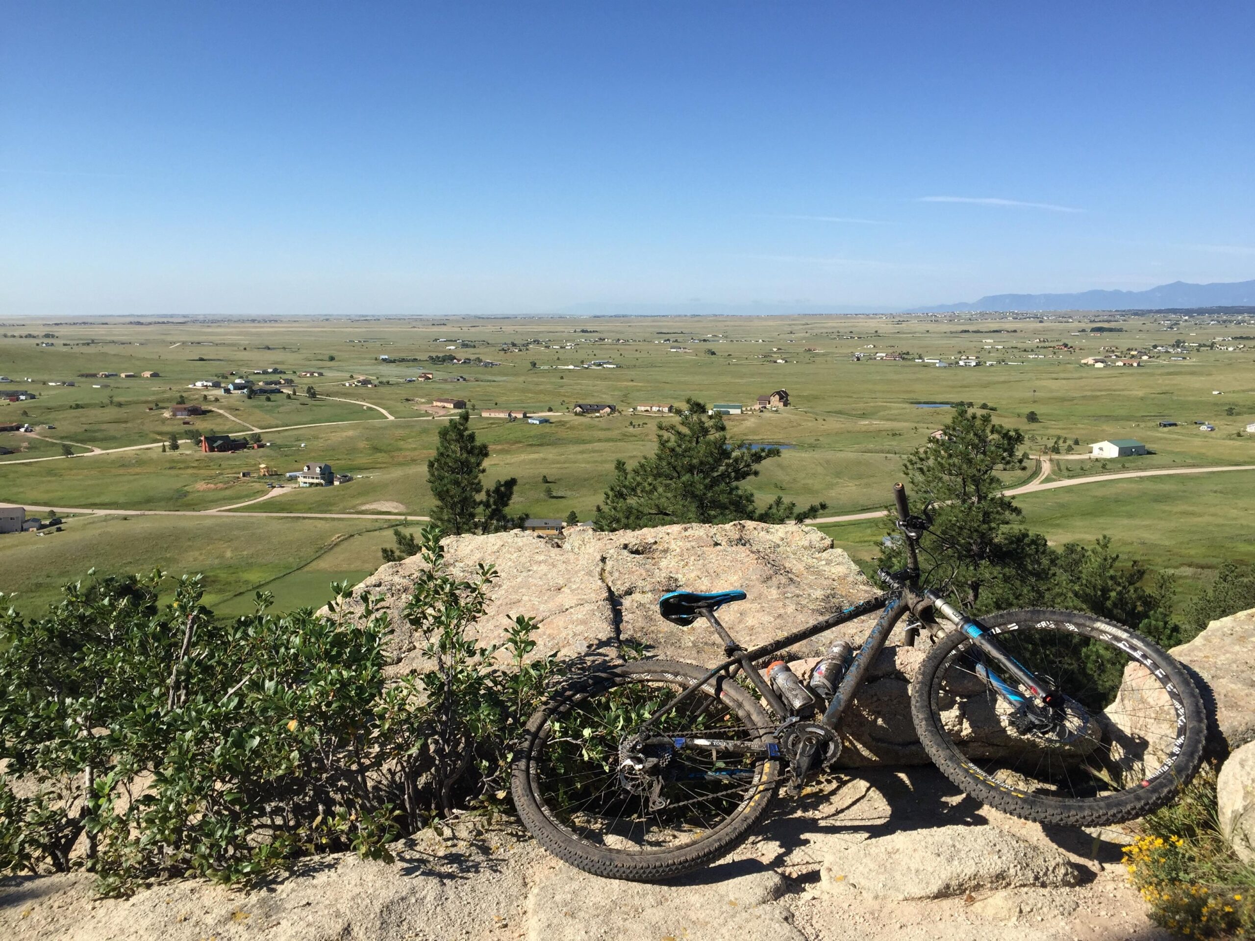 A mountain bike resting on a rocky ledge with a panoramic view of a rural landscape below. The scene features rolling green hills dotted with houses and winding dirt roads, under a clear blue sky. Homestead Ranch Regional Park mountain bike trail.