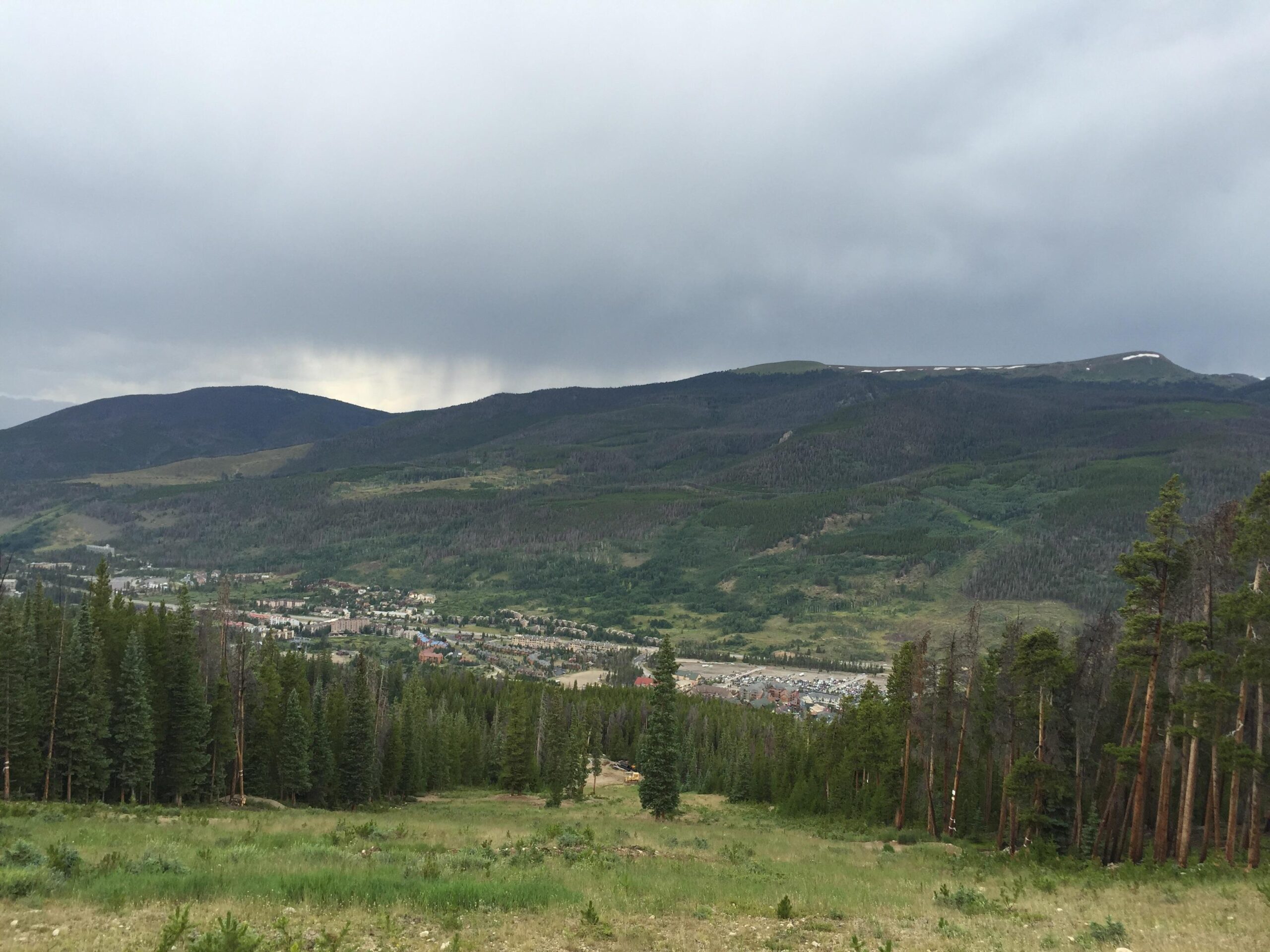 A panoramic view of a mountainous landscape under a cloudy sky. The foreground features a mix of green grass and pine trees, while the midground showcases a small town nestled in a valley surrounded by rolling hills and forested areas. The distant mountains rise prominently, with hints of snow visible on their peaks, suggesting a cool, overcast day. Keystone Resort Bike Park mountain bike trail.