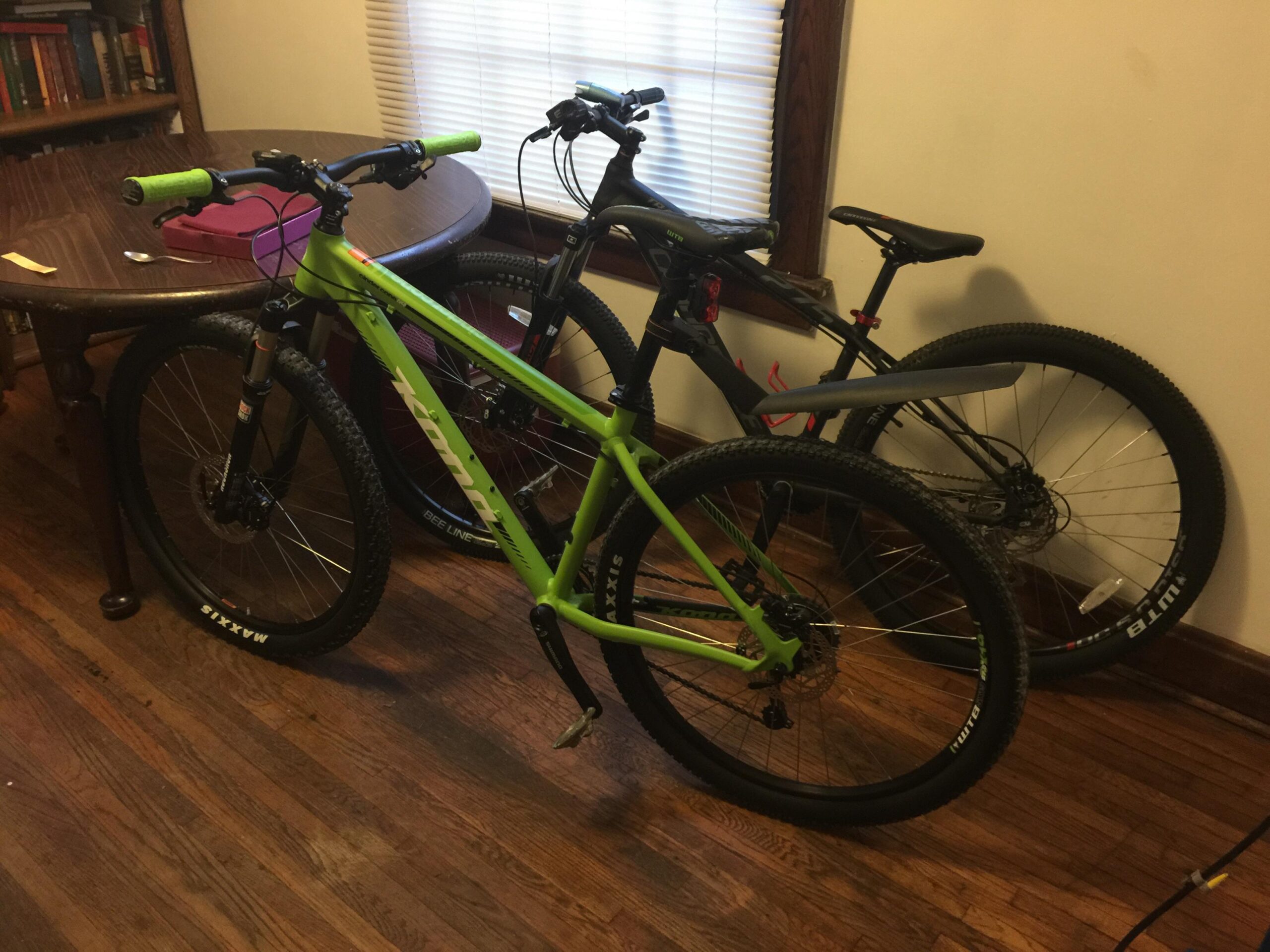 Kona Cinder Cone: Two bicycles are parked inside a room with wooden floors. The first bicycle is bright green with black components and features knobby tires, while the second bicycle is black with a more subdued design and similar tire styling. A round wooden table is visible in the background, along with a few books on a shelf and a window with blinds. The setting suggests a casual indoor space.