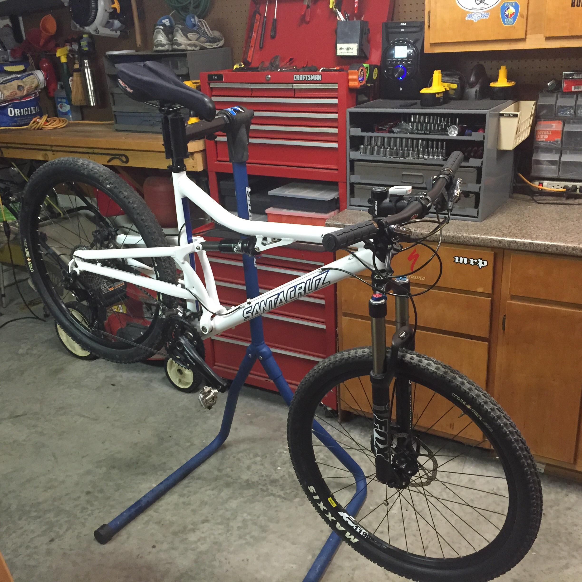 Santa Cruz Superlight: A white mountain bike is mounted on a repair stand in a garage workspace, surrounded by tools and storage boxes. The background features a red toolbox, shelves with assorted tools, and some equipment hanging on the walls. The bike's front wheel is off the ground, and the focus is on the bike's frame and suspension.