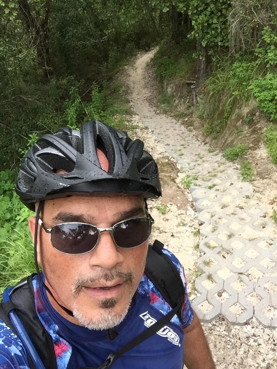 A person wearing a helmet and sunglasses takes a selfie while standing next to a narrow, winding bike trail surrounded by lush greenery. The path is made of interlocking stones and disappears into the dense foliage in the background. Balm Boyette Scrub Preserve mountain bike trail.