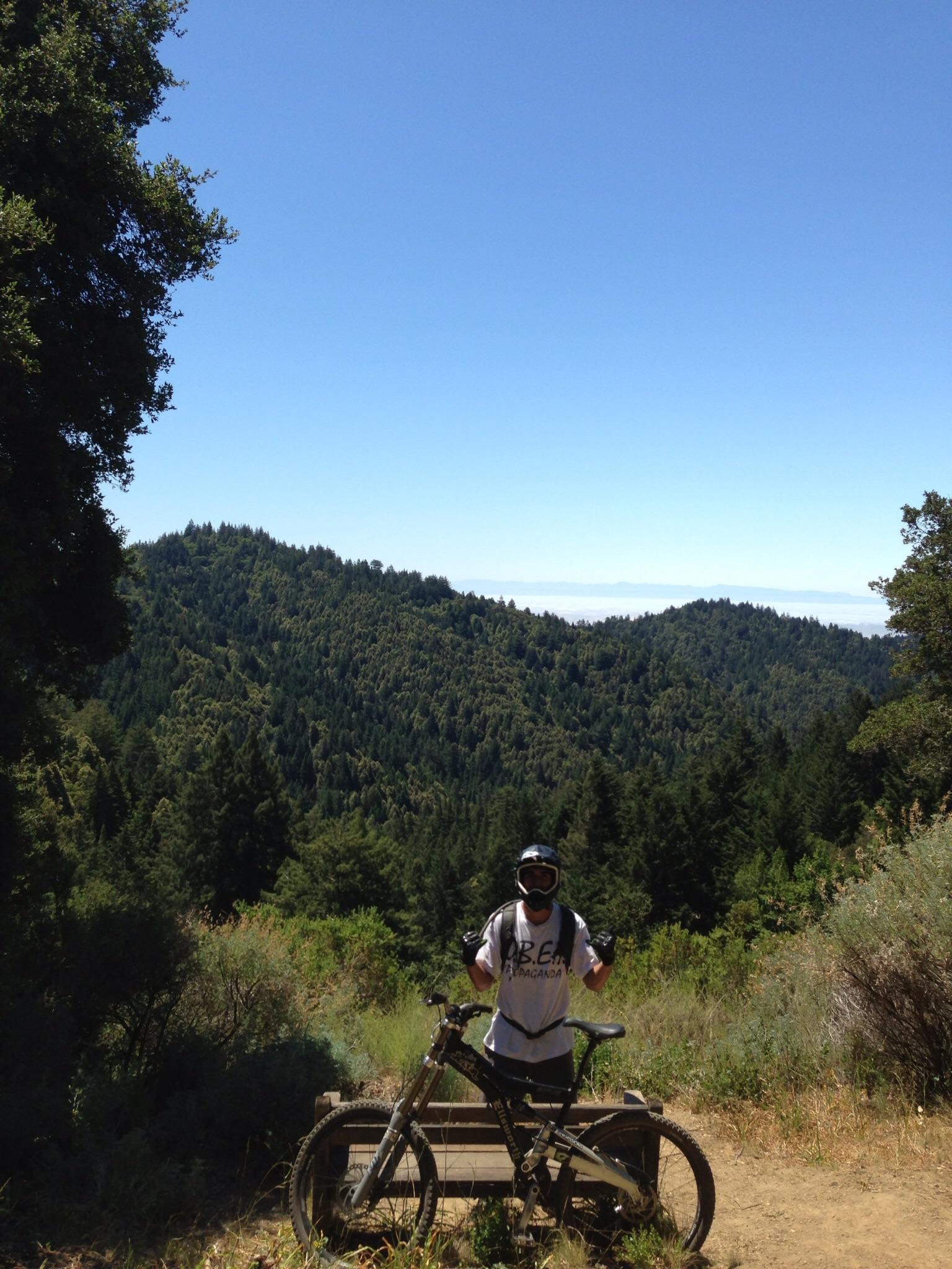A person wearing a helmet and protective gear stands next to a mountain bike, giving a thumbs-up in a scenic outdoor setting. In the background, lush green hills and a clear blue sky create a picturesque landscape. Forest Of Nisene Marks and Soquel Demonstration Forest mountain bike trail.