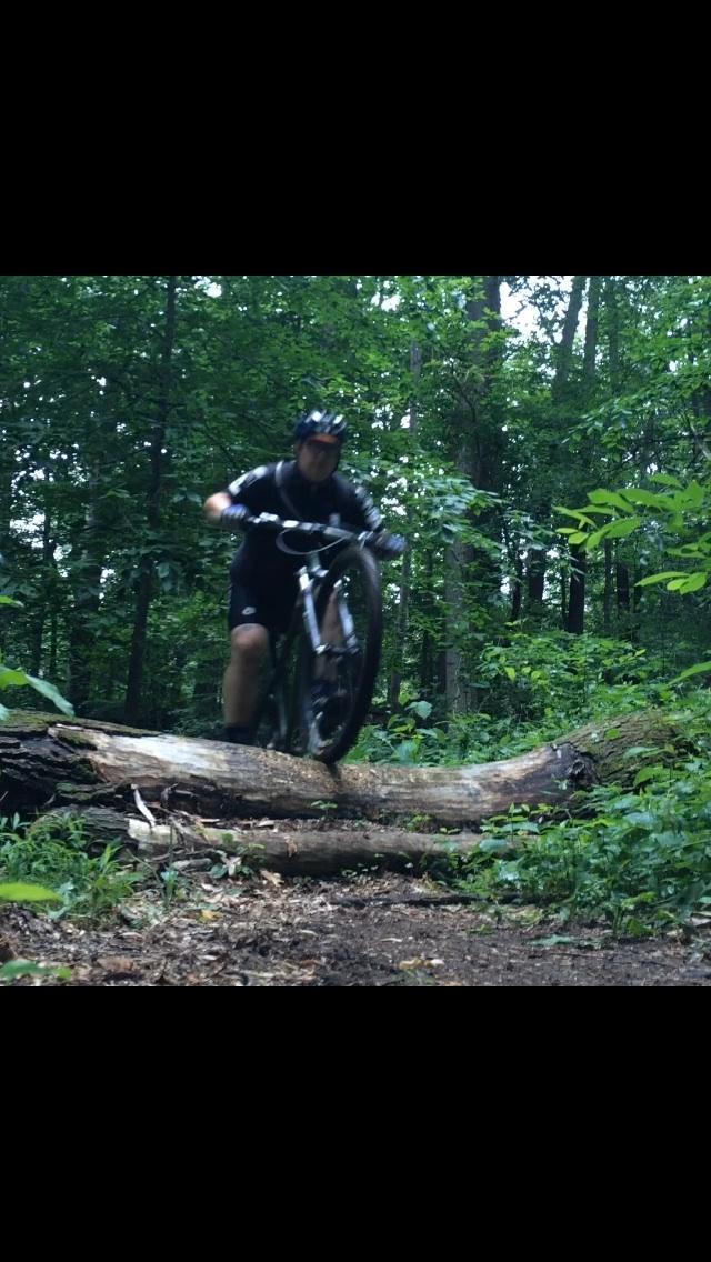A mountain biker performing a jump over a fallen log on a wooded trail, surrounded by green foliage and trees. The rider is wearing a helmet and biking gear, with one wheel lifted off the ground. Brandywine State Park mountain bike trail.